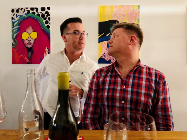 Two men sitting at a table in a gallery with wine bottles in front of them.
